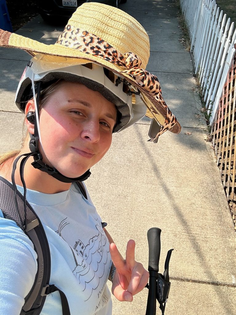 Una mujer con casco y sombrero, montando en bicicleta, haciendo el signo de la paz con la mano y sonriendo.