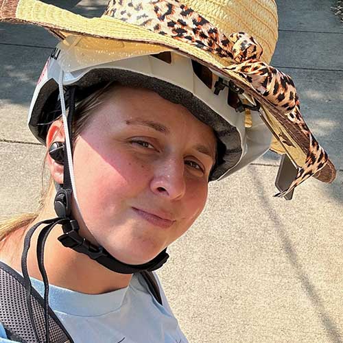 A woman wearing a helmet and a hat riding a bike doing a peace sign with her hand and smiling.